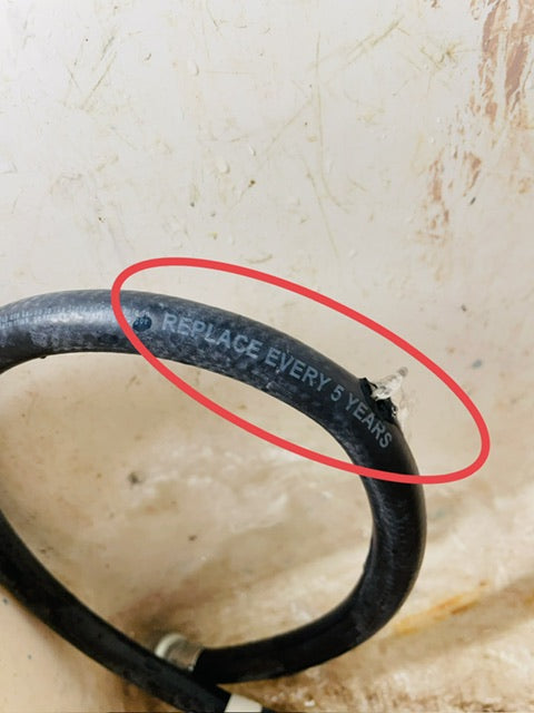 A black rubber washing machine hose with water spewing out of a section that has burst. The water burst occurred right next to the text that says “replace every 5 years.” This is a common occurrence when washing machine hoses are not replaced regularly.